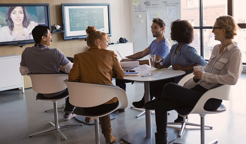 Group of people in a room in a virtual meeting with another woman.