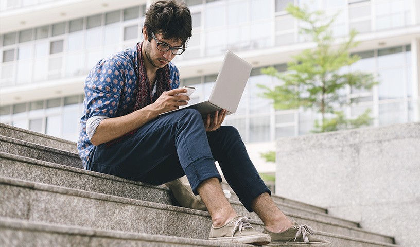 A man sitting on steps outside of a building looking at a mobile phone in one hand while holding a laptop in his other hand.