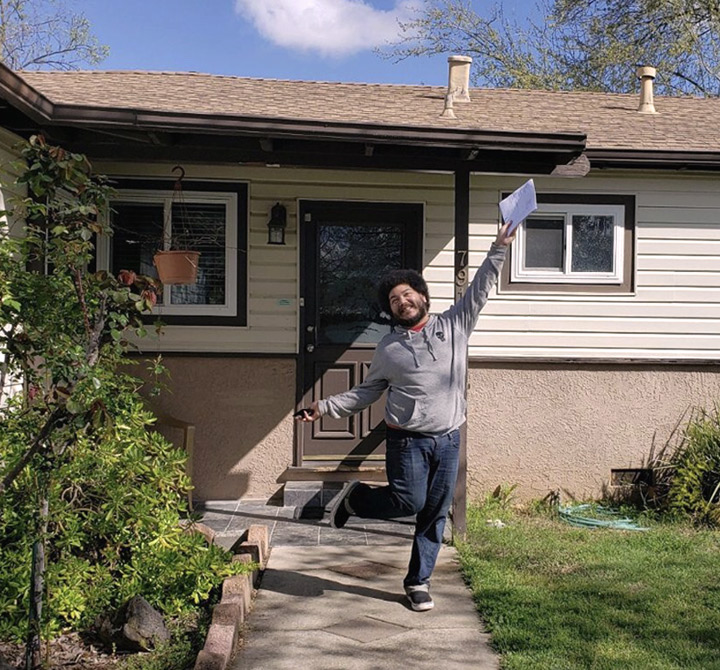 A happy young man is standing outside on one leg with one arm in the air holding a document in front of a house.